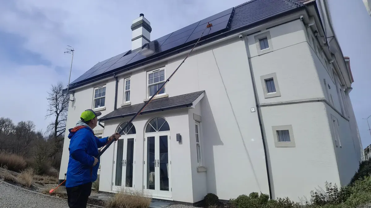 A cleaner using a long pole to clean the solar panels on the roof of a white house